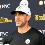 Aaron Rodgers in einem schwarzen Hemd mit einem Steelers-Logo und einer weißen Steelers-Mütze lächelt, während er auf einer Presseveranstaltung über die bevorstehende NFL-Saison 2025 spricht. Hinter ihm ist ein Hintergrund mit dem Steelers- und dem PNC-Logo zu sehen, und vor ihm befinden sich mehrere Mikrofone. Diese Beschreibung wurde mit der FootballR KI automatisch generiert.