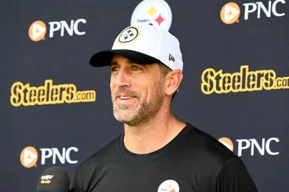 Aaron Rodgers in einem schwarzen Hemd mit einem Steelers-Logo und einer weißen Steelers-Mütze lächelt, während er auf einer Presseveranstaltung über die bevorstehende NFL-Saison 2025 spricht. Hinter ihm ist ein Hintergrund mit dem Steelers- und dem PNC-Logo zu sehen, und vor ihm befinden sich mehrere Mikrofone. Diese Beschreibung wurde mit der FootballR KI automatisch generiert.
