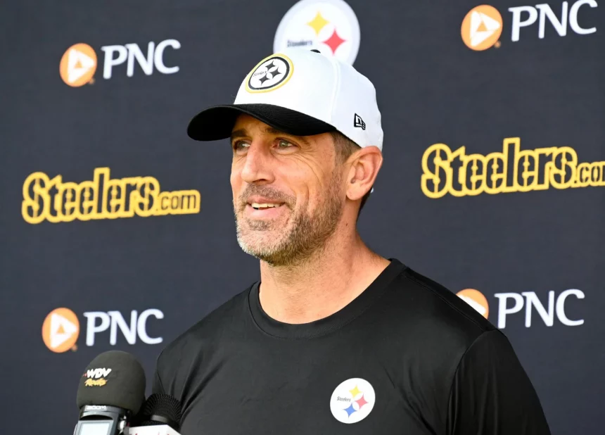 Aaron Rodgers in einem schwarzen Hemd mit einem Steelers-Logo und einer weißen Steelers-Mütze lächelt, während er auf einer Presseveranstaltung über die bevorstehende NFL-Saison 2025 spricht. Hinter ihm ist ein Hintergrund mit dem Steelers- und dem PNC-Logo zu sehen, und vor ihm befinden sich mehrere Mikrofone. Diese Beschreibung wurde mit der FootballR KI automatisch generiert.
