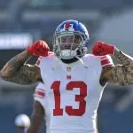 Odell Beckham Jr., in einem weißen Trikot der New York Giants mit der Nummer 13, lässt auf dem Spielfeld seine Muskeln spielen. Er trägt einen blauen Helm, rote Handschuhe und Tattoos auf beiden Armen. Der unscharfe Stadionhintergrund zeigt leere Sitze. Diese Beschreibung wurde mit der FootballR KI automatisch generiert.