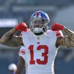 Odell Beckham Jr., in einem weißen Trikot der New York Giants mit der Nummer 13, lässt auf dem Spielfeld seine Muskeln spielen. Er trägt einen blauen Helm, rote Handschuhe und Tattoos auf beiden Armen. Der unscharfe Stadionhintergrund zeigt leere Sitze. Diese Beschreibung wurde mit der FootballR KI automatisch generiert.