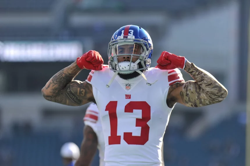 Odell Beckham Jr., in einem weißen Trikot der New York Giants mit der Nummer 13, lässt auf dem Spielfeld seine Muskeln spielen. Er trägt einen blauen Helm, rote Handschuhe und Tattoos auf beiden Armen. Der unscharfe Stadionhintergrund zeigt leere Sitze. Diese Beschreibung wurde mit der FootballR KI automatisch generiert.