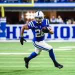 Ein Footballspieler in einem blau-weißen Trikot der Indianapolis Colts, der den Ball hält, läuft während der NFL Preseason auf einem grünen Feld nach vorne. Er weicht einem Verteidiger in Weiß aus, der sich von links nähert, wobei er ein Bein in der Luft hat und Helm und Handschuhe trägt. Diese Beschreibung wurde mit der FootballR KI automatisch generiert.