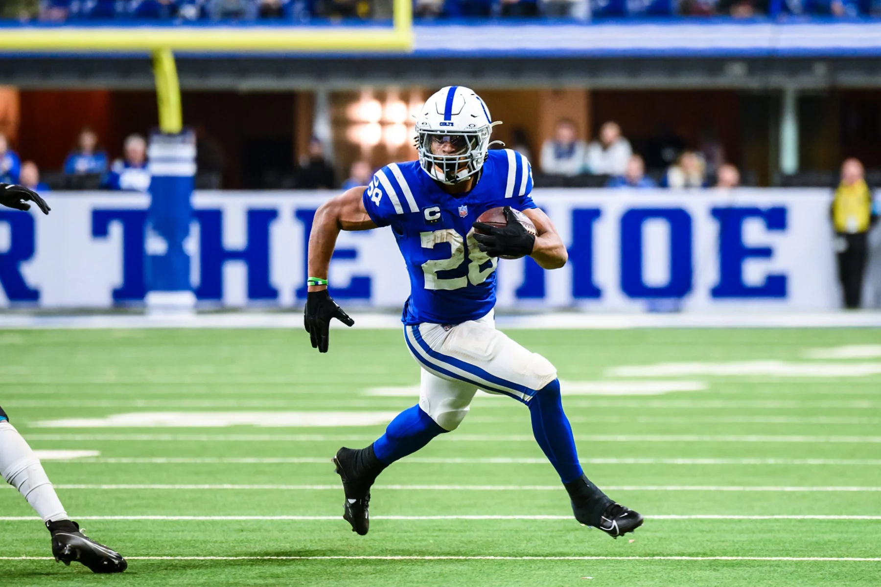 Ein Footballspieler in einem blau-weißen Trikot der Indianapolis Colts, der den Ball hält, läuft während der NFL Preseason auf einem grünen Feld nach vorne. Er weicht einem Verteidiger in Weiß aus, der sich von links nähert, wobei er ein Bein in der Luft hat und Helm und Handschuhe trägt. Diese Beschreibung wurde mit der FootballR KI automatisch generiert.