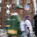 Cap Space: Menschen gehen am Stadion von Tottenham Hotspur vorbei, über dem Schilder für Tottenham Hotspur, NFL und F1 Drive London zu sehen sind. Einige Menschen in NFL-Trikots diskutieren über die Spielfeldbegrenzung, während sich die verschwommene Menge an der modernen, holzgetäfelten Fassade des Gebäudes vorbeischiebt. Diese Beschreibung wurde mit der FootballR KI automatisch generiert.