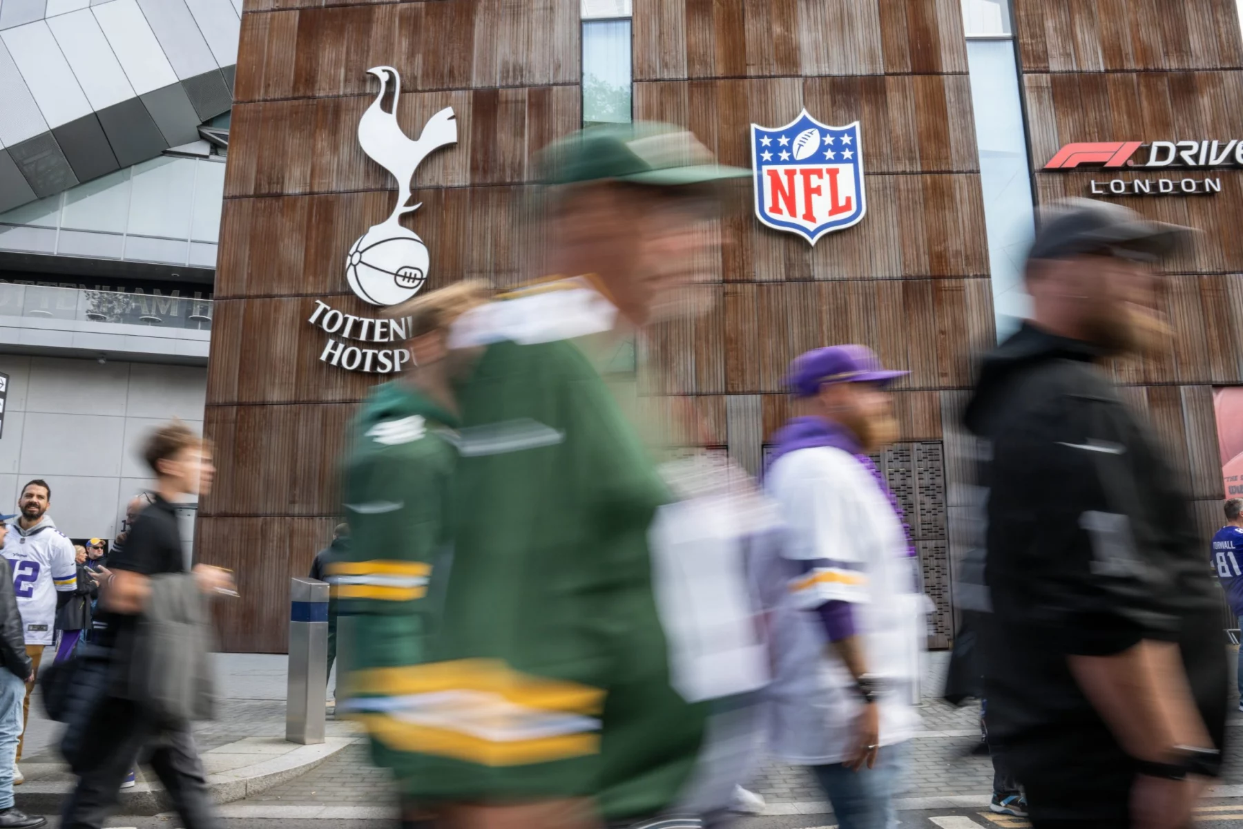 Cap Space: Menschen gehen am Stadion von Tottenham Hotspur vorbei, über dem Schilder für Tottenham Hotspur, NFL und F1 Drive London zu sehen sind. Einige Menschen in NFL-Trikots diskutieren über die Spielfeldbegrenzung, während sich die verschwommene Menge an der modernen, holzgetäfelten Fassade des Gebäudes vorbeischiebt. Diese Beschreibung wurde mit der FootballR KI automatisch generiert.