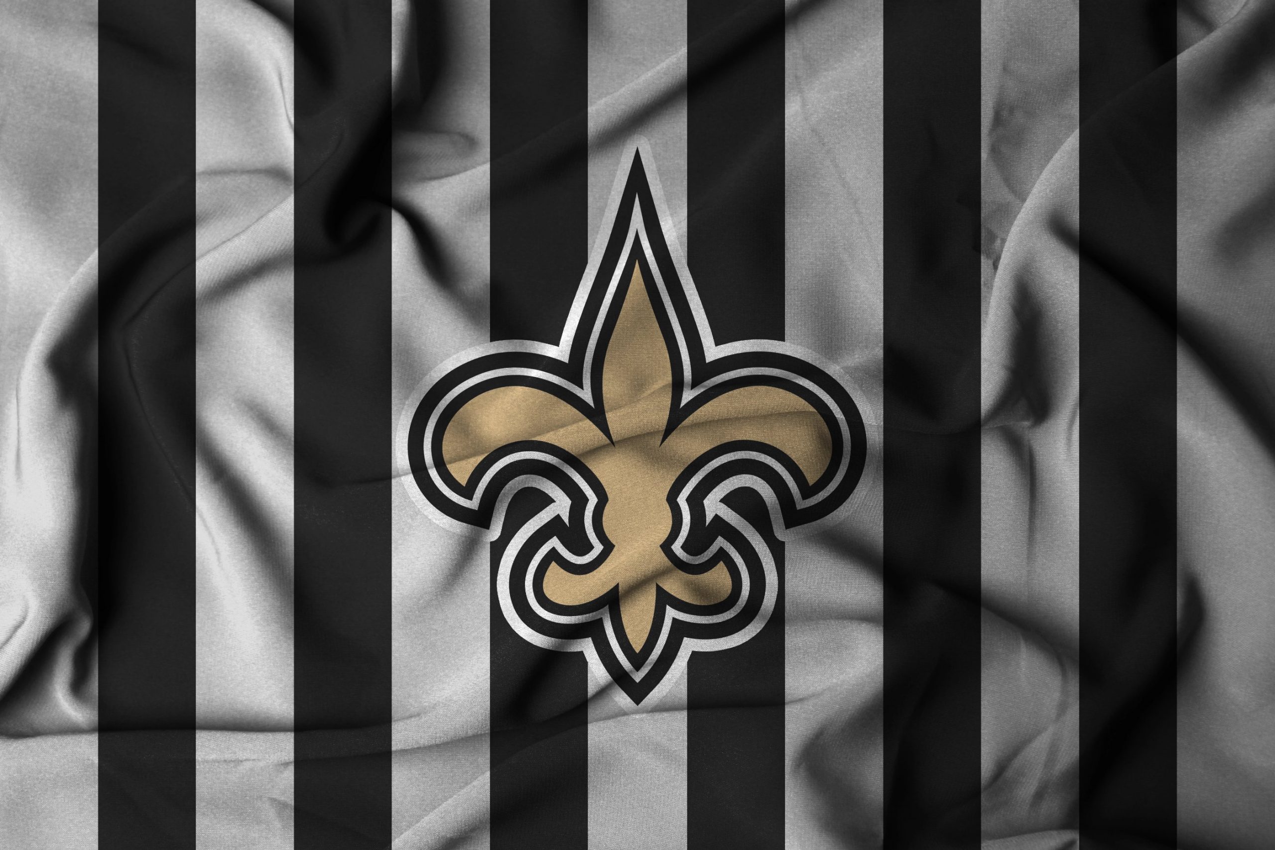 New Orleans Saints - Ein zerknitterter Stoff mit abwechselnd schwarzen und grauen vertikalen Streifen. In der Mitte befindet sich eine große goldene Fleur-de-Lis, das ikonische Logo der New Orleans Saints. Die Falten des Stoffes erzeugen Schatten und Textur, die das Design der neuen Farbe noch besser zur Geltung bringen. Diese Beschreibung wurde mit der FootballR KI automatisch generiert.
