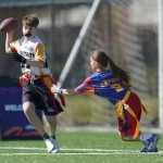 Ein Junge in einem weiß-orangenen Trikot rennt mit einem Football, während ein Mädchen in einer blau-roten Uniform nach seiner Flagge greift. Beide spielen NFL Flag Tournament auf einem grünen Feld, mit einem unscharfen Sportnetz und einem Schild im Hintergrund. Diese Beschreibung wurde mit der FootballR KI automatisch generiert.