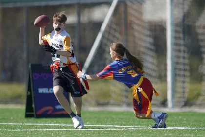 Ein Junge in einem weiß-orangenen Trikot rennt mit einem Football, während ein Mädchen in einer blau-roten Uniform nach seiner Flagge greift. Beide spielen NFL Flag Tournament auf einem grünen Feld, mit einem unscharfen Sportnetz und einem Schild im Hintergrund. Diese Beschreibung wurde mit der FootballR KI automatisch generiert.