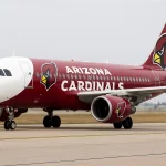 Ein kastanienbraunes und weißes Flugzeug mit dem Schriftzug "Arizona Cardinals", dem Vogellogo des Teams und dem Branding von Cardinals Premier Travel rollt auf der Landebahn. Die Nase ist weiß, während sich der kastanienbraune Rumpf vom Gras, der Landebahn und dem bewölkten Himmel abhebt. Diese Beschreibung wurde mit der FootballR KI automatisch generiert.