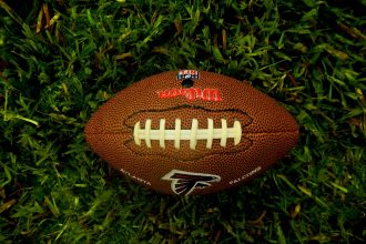 Ein brauner Wilson NFL-Football mit weißen Schnürsenkeln und dem Logo der Atlanta Falcons ruht auf grünem Gras. Die Textur des Balles und die einzelnen Grashalme sind deutlich zu erkennen. Von oben aufgenommen, unterstreicht er die Begeisterung der NFL-Fans in Österreich und Wettgiganten gleichermaßen. Diese Beschreibung wurde mit der FootballR KI automatisch generiert.