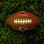Ein brauner Wilson NFL-Football mit weißen Schnürsenkeln und dem Logo der Atlanta Falcons ruht auf grünem Gras. Die Textur des Balles und die einzelnen Grashalme sind deutlich zu erkennen. Von oben aufgenommen, unterstreicht er die Begeisterung der NFL-Fans in Österreich und Wettgiganten gleichermaßen. Diese Beschreibung wurde mit der FootballR KI automatisch generiert.