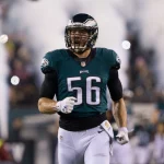 Ein Footballspieler, Bryan Braman, trägt ein grünes Philadelphia Eagles-Trikot mit der Nummer 56 und läuft lächelnd auf das Spielfeld. Rauch und verschwommene Mannschaftskameraden im Hintergrund deuten auf einen dramatischen Auftritt oder eine Atmosphäre vor dem Spiel hin. Diese Beschreibung wurde mit der FootballR KI automatisch generiert.