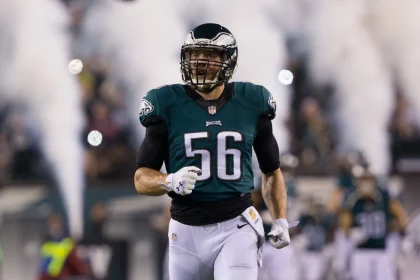 Ein Footballspieler, Bryan Braman, trägt ein grünes Philadelphia Eagles-Trikot mit der Nummer 56 und läuft lächelnd auf das Spielfeld. Rauch und verschwommene Mannschaftskameraden im Hintergrund deuten auf einen dramatischen Auftritt oder eine Atmosphäre vor dem Spiel hin. Diese Beschreibung wurde mit der FootballR KI automatisch generiert.