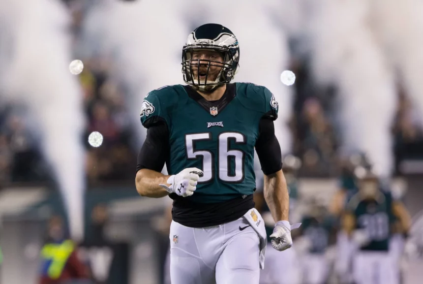 Ein Footballspieler, Bryan Braman, trägt ein grünes Philadelphia Eagles-Trikot mit der Nummer 56 und läuft lächelnd auf das Spielfeld. Rauch und verschwommene Mannschaftskameraden im Hintergrund deuten auf einen dramatischen Auftritt oder eine Atmosphäre vor dem Spiel hin. Diese Beschreibung wurde mit der FootballR KI automatisch generiert.