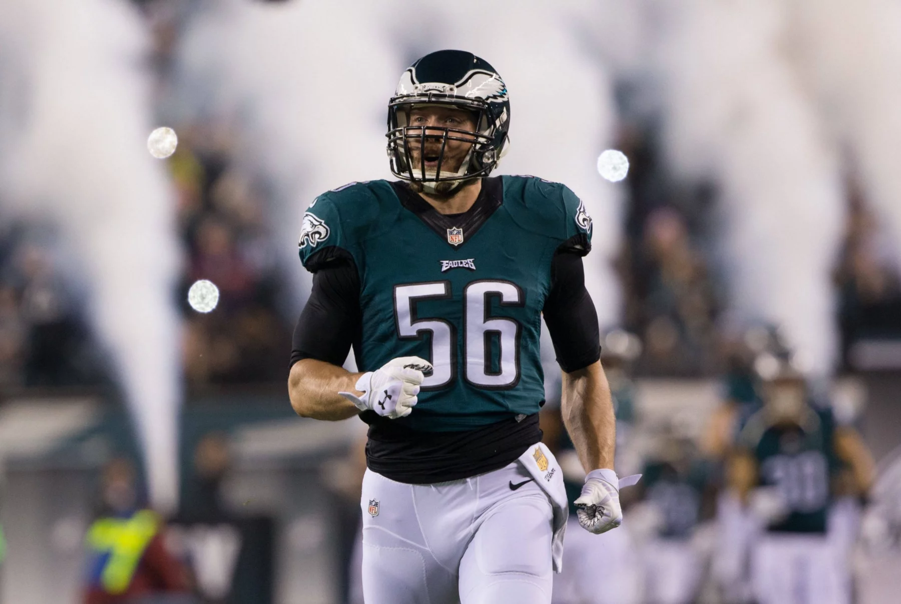 Ein Footballspieler, Bryan Braman, trägt ein grünes Philadelphia Eagles-Trikot mit der Nummer 56 und läuft lächelnd auf das Spielfeld. Rauch und verschwommene Mannschaftskameraden im Hintergrund deuten auf einen dramatischen Auftritt oder eine Atmosphäre vor dem Spiel hin. Diese Beschreibung wurde mit der FootballR KI automatisch generiert.