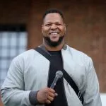 Ndamukong Suh steht im Freien, lächelt und hält ein Mikrofon in der Hand. Er trägt eine hellgraue Jacke über einem dunklen Hemd, was an den lässigen Stil von Ndamukong Suh erinnert. Im Hintergrund sind eine Backsteinmauer und ein großes Fenster mit einem Gittermuster zu sehen. Diese Beschreibung wurde mit der FootballR KI automatisch generiert.