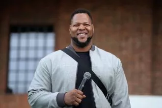 Ndamukong Suh steht im Freien, lächelt und hält ein Mikrofon in der Hand. Er trägt eine hellgraue Jacke über einem dunklen Hemd, was an den lässigen Stil von Ndamukong Suh erinnert. Im Hintergrund sind eine Backsteinmauer und ein großes Fenster mit einem Gittermuster zu sehen. Diese Beschreibung wurde mit der FootballR KI automatisch generiert.