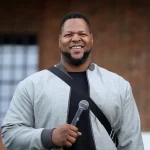 Ndamukong Suh steht im Freien, lächelt und hält ein Mikrofon in der Hand. Er trägt eine hellgraue Jacke über einem dunklen Hemd, was an den lässigen Stil von Ndamukong Suh erinnert. Im Hintergrund sind eine Backsteinmauer und ein großes Fenster mit einem Gittermuster zu sehen. Diese Beschreibung wurde mit der FootballR KI automatisch generiert.