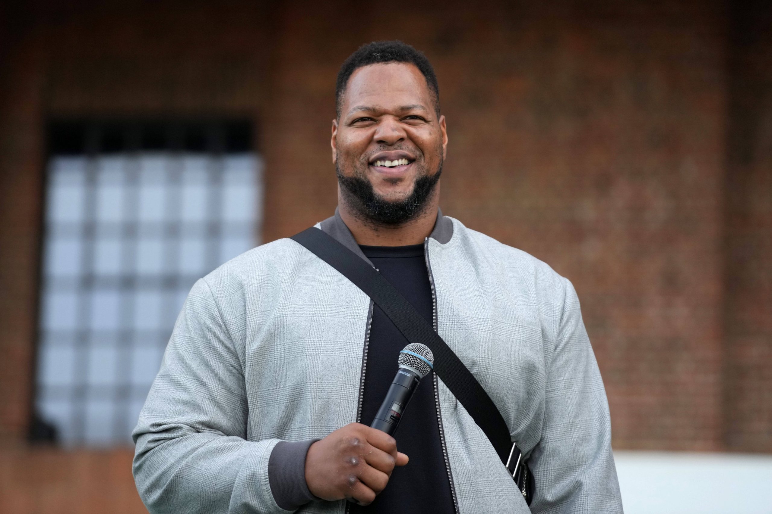 Ndamukong Suh steht im Freien, lächelt und hält ein Mikrofon in der Hand. Er trägt eine hellgraue Jacke über einem dunklen Hemd, was an den lässigen Stil von Ndamukong Suh erinnert. Im Hintergrund sind eine Backsteinmauer und ein großes Fenster mit einem Gittermuster zu sehen. Diese Beschreibung wurde mit der FootballR KI automatisch generiert.