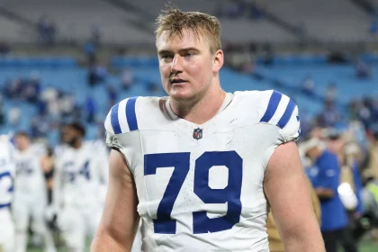 Der Footballspieler Bernhard Raimann, der ein weißes Trikot mit blauen Streifen und der Nummer 79 trägt, betritt das Spielfeld. Er wirkt verschwitzt und konzentriert, hat kurzes blondes Haar. Im Hintergrund sind andere Spieler und eine unscharfe Stadionbestuhlung zu sehen. Diese Beschreibung wurde mit der FootballR KI automatisch generiert.