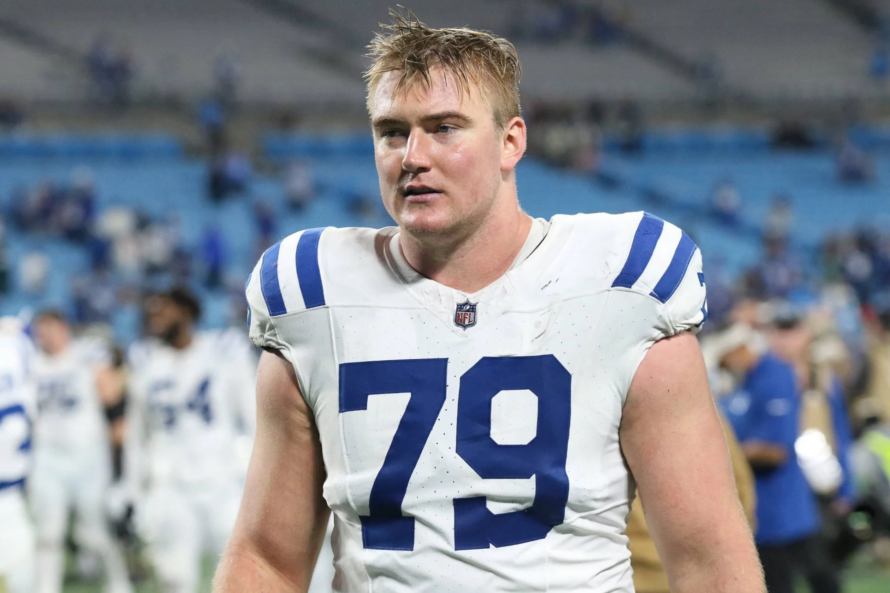 Der Footballspieler Bernhard Raimann, der ein weißes Trikot mit blauen Streifen und der Nummer 79 trägt, betritt das Spielfeld. Er wirkt verschwitzt und konzentriert, hat kurzes blondes Haar. Im Hintergrund sind andere Spieler und eine unscharfe Stadionbestuhlung zu sehen. Diese Beschreibung wurde mit der FootballR KI automatisch generiert.