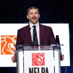 JC Tretter, in einem kastanienbraunen Anzug und mit schwarzer Krawatte, spricht an einem durchsichtigen Podium mit einem NFLPA-Logo. Hinter ihm zeigt ein Bildschirm dasselbe Logo mit einem stilisierten Football-Spieler in Weiß und Orange vor dunkelroter und blauer Hintergrundbeleuchtung. Diese Beschreibung wurde mit der FootballR KI automatisch generiert.