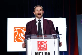 JC Tretter, in einem kastanienbraunen Anzug und mit schwarzer Krawatte, spricht an einem durchsichtigen Podium mit einem NFLPA-Logo. Hinter ihm zeigt ein Bildschirm dasselbe Logo mit einem stilisierten Football-Spieler in Weiß und Orange vor dunkelroter und blauer Hintergrundbeleuchtung. Diese Beschreibung wurde mit der FootballR KI automatisch generiert.