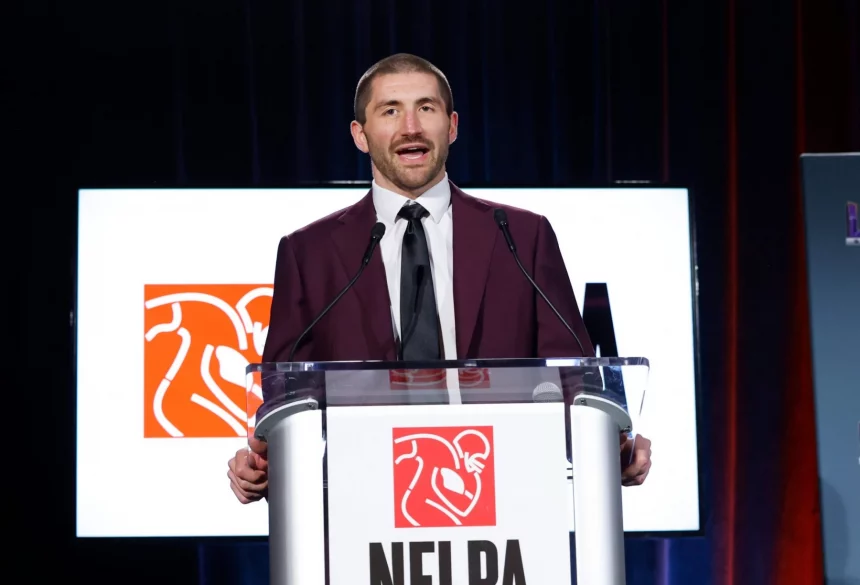 JC Tretter, in einem kastanienbraunen Anzug und mit schwarzer Krawatte, spricht an einem durchsichtigen Podium mit einem NFLPA-Logo. Hinter ihm zeigt ein Bildschirm dasselbe Logo mit einem stilisierten Football-Spieler in Weiß und Orange vor dunkelroter und blauer Hintergrundbeleuchtung. Diese Beschreibung wurde mit der FootballR KI automatisch generiert.