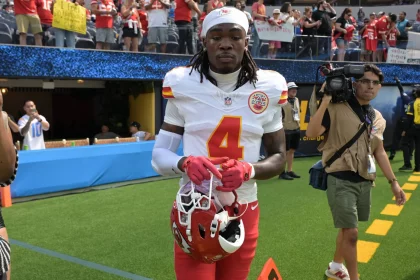 Rashee Rice in der Uniform der Kansas City Chiefs, der die Nummer 4 trägt, läuft mit seinem Helm auf das Spielfeld. Rashee Rice erscheint konzentriert, mit langen Haaren und einem weißen Stirnband, während Fans in Rot die Tribüne füllen und ein Kameramann in der Nähe aufnimmt. Diese Beschreibung wurde mit der FootballR KI automatisch generiert.