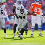 Ein Football-Spieler, Christian Wilkins, in einem weißen Las Vegas Raiders-Trikot (#94) breitet seine Arme zum Jubel auf dem Spielfeld aus, umgeben von Mannschaftskameraden und Denver Broncos-Spielern in orangefarbenen Uniformen; Sonnenlicht beleuchtet die Aktion. Diese Beschreibung wurde mit der FootballR KI automatisch generiert.