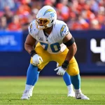 Rashawn Slater, die Nummer 70 der Los Angeles Chargers, hockt in seinem weiß-gelben Trikot auf dem Spielfeld und hält sich bereit. Er trägt einen Helm mit dem Blitz-Logo, während im Hintergrund verschwommene Zuschauer und eine Anzeigetafel mit der Aufschrift "4:0" zu sehen sind. Diese Beschreibung wurde mit der FootballR KI automatisch generiert.