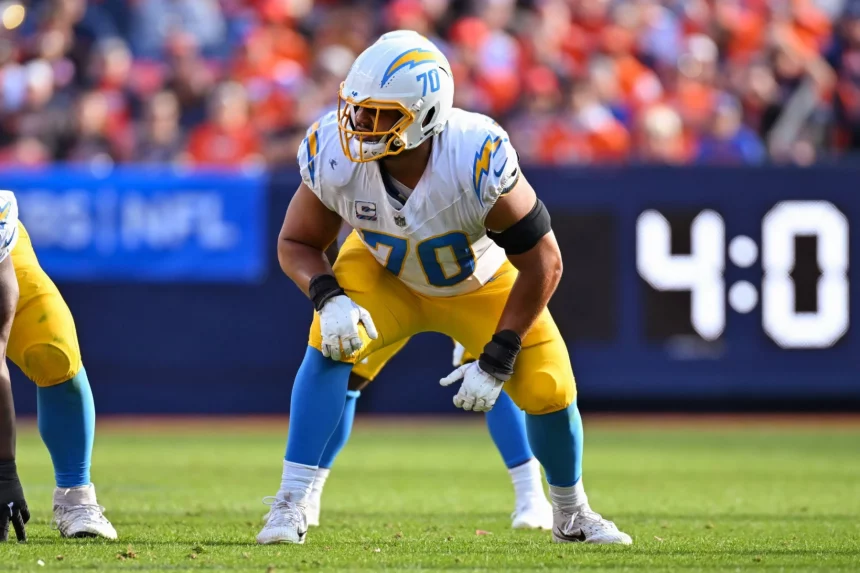 Rashawn Slater, die Nummer 70 der Los Angeles Chargers, hockt in seinem weiß-gelben Trikot auf dem Spielfeld und hält sich bereit. Er trägt einen Helm mit dem Blitz-Logo, während im Hintergrund verschwommene Zuschauer und eine Anzeigetafel mit der Aufschrift "4:0" zu sehen sind. Diese Beschreibung wurde mit der FootballR KI automatisch generiert.