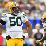 Ein Footballspieler der Green Bay Packers, Zach Tom, trägt ein weißes Trikot mit der Nummer 50, eine gelbe Hose und einen gelben Helm und steht während eines Spiels mit den Händen in den Hüften. Teamkollegen in ähnlichen Trikots sind im Hintergrund auf dem Spielfeld zu sehen. Diese Beschreibung wurde mit der FootballR KI automatisch generiert.