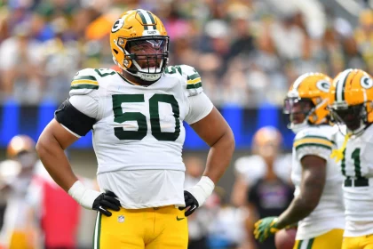 Ein Footballspieler der Green Bay Packers, Zach Tom, trägt ein weißes Trikot mit der Nummer 50, eine gelbe Hose und einen gelben Helm und steht während eines Spiels mit den Händen in den Hüften. Teamkollegen in ähnlichen Trikots sind im Hintergrund auf dem Spielfeld zu sehen. Diese Beschreibung wurde mit der FootballR KI automatisch generiert.