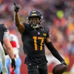 Ein Football-Spieler in einem schwarzen Trikot mit der Nummer 17 und einem goldenen "W" auf dem Helm - Terry McLaurin - jubelt auf dem Spielfeld, zeigt mit der rechten Hand nach oben und hält einen Football in der linken Hand. Ein anderer Spieler in einer weiß-blauen Uniform mit der Nummer 44 steht in der Nähe. Diese Beschreibung wurde mit der FootballR KI automatisch generiert.