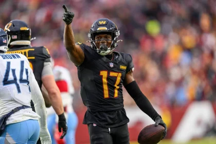 Ein Football-Spieler in einem schwarzen Trikot mit der Nummer 17 und einem goldenen "W" auf dem Helm - Terry McLaurin - jubelt auf dem Spielfeld, zeigt mit der rechten Hand nach oben und hält einen Football in der linken Hand. Ein anderer Spieler in einer weiß-blauen Uniform mit der Nummer 44 steht in der Nähe. Diese Beschreibung wurde mit der FootballR KI automatisch generiert.