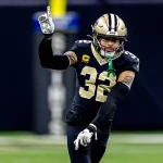 Ein Football-Spieler, identifiziert als Tyrann Mathieu, in einer schwarzen New Orleans Saints-Uniform mit der Nummer 32 und einem Kapitänsaufnäher, gestikuliert mit der erhobenen rechten Hand. Er scheint auf dem Spielfeld ein Zeichen zu geben, sein goldener Helm leuchtet vor dem unscharfen Hintergrund. Diese Beschreibung wurde mit der FootballR KI automatisch generiert.