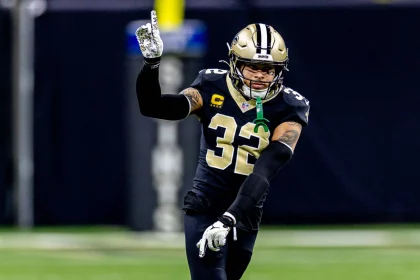 Ein Football-Spieler, identifiziert als Tyrann Mathieu, in einer schwarzen New Orleans Saints-Uniform mit der Nummer 32 und einem Kapitänsaufnäher, gestikuliert mit der erhobenen rechten Hand. Er scheint auf dem Spielfeld ein Zeichen zu geben, sein goldener Helm leuchtet vor dem unscharfen Hintergrund. Diese Beschreibung wurde mit der FootballR KI automatisch generiert.