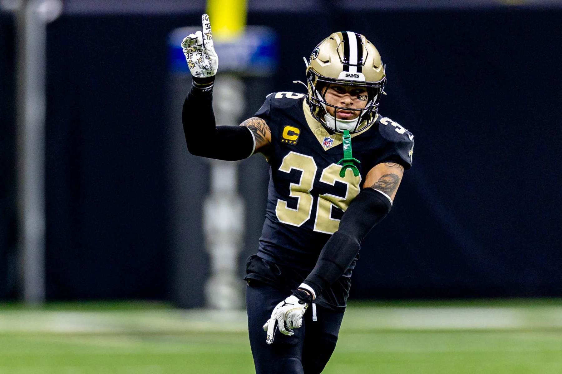 Ein Football-Spieler, identifiziert als Tyrann Mathieu, in einer schwarzen New Orleans Saints-Uniform mit der Nummer 32 und einem Kapitänsaufnäher, gestikuliert mit der erhobenen rechten Hand. Er scheint auf dem Spielfeld ein Zeichen zu geben, sein goldener Helm leuchtet vor dem unscharfen Hintergrund. Diese Beschreibung wurde mit der FootballR KI automatisch generiert.