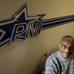 Ein Mann mit kurzen grauen Haaren, Brille und gestreiftem Pullover sitzt lächelnd neben einer Wand mit dem fetten Schriftzug "PNI" und einem großen blau-weißen Sternlogo - ein Hinweis auf Pettis Norman -, während in der unteren Ecke teilweise Blattpflanzen zu sehen sind. Diese Beschreibung wurde mit der FootballR KI automatisch generiert.