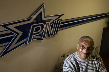Ein Mann mit kurzen grauen Haaren, Brille und gestreiftem Pullover sitzt lächelnd neben einer Wand mit dem fetten Schriftzug "PNI" und einem großen blau-weißen Sternlogo - ein Hinweis auf Pettis Norman -, während in der unteren Ecke teilweise Blattpflanzen zu sehen sind. Diese Beschreibung wurde mit der FootballR KI automatisch generiert.