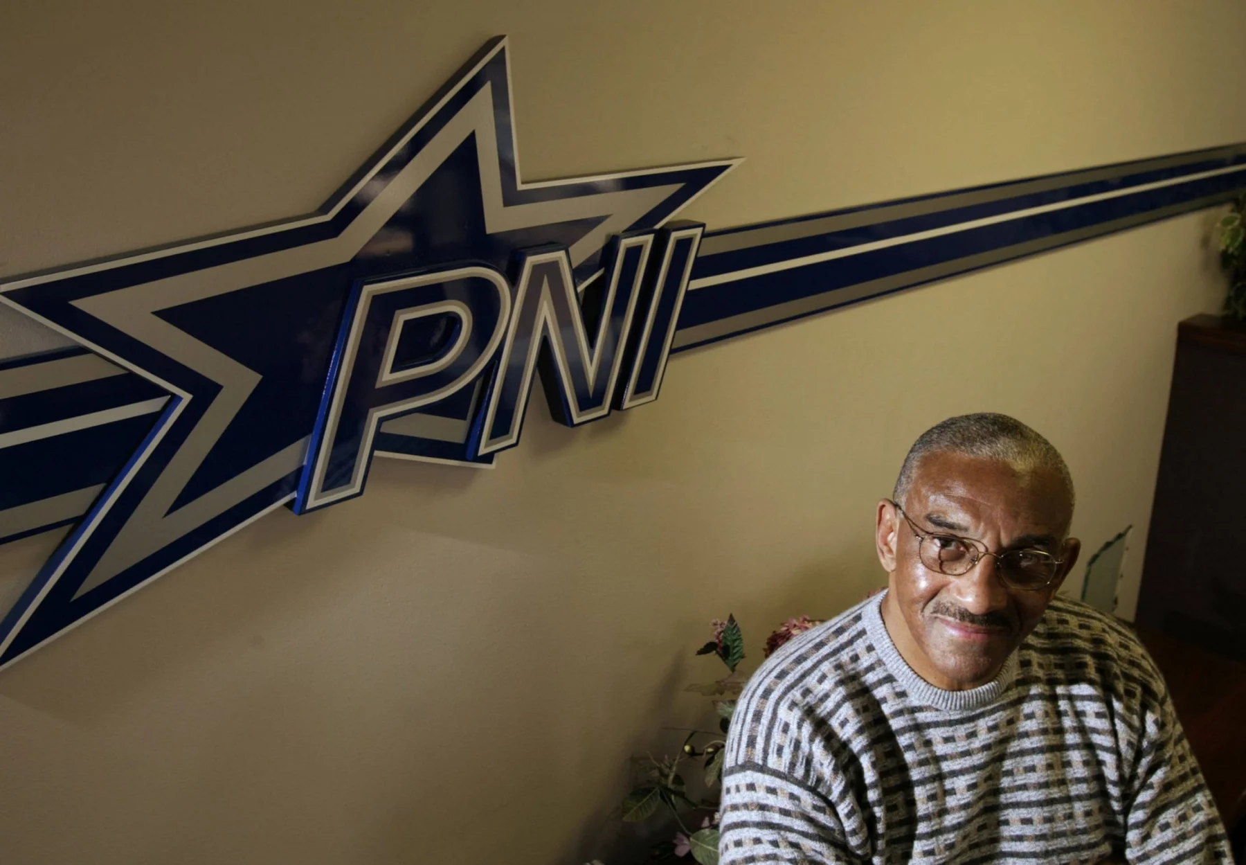 Ein Mann mit kurzen grauen Haaren, Brille und gestreiftem Pullover sitzt lächelnd neben einer Wand mit dem fetten Schriftzug "PNI" und einem großen blau-weißen Sternlogo - ein Hinweis auf Pettis Norman -, während in der unteren Ecke teilweise Blattpflanzen zu sehen sind. Diese Beschreibung wurde mit der FootballR KI automatisch generiert.