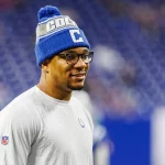 Ein lächelnder Anthony Richardson in einem weißen Hemd mit Colts-Logo und einer blauen Mütze mit der Aufschrift "COLTS" steht auf dem Spielfeld. Er trägt eine Brille und ist scharf eingestellt, während das Stadion hinter ihm nach einem spannenden Quarterback-Duell verschwimmt. Diese Beschreibung wurde mit der FootballR KI automatisch generiert.