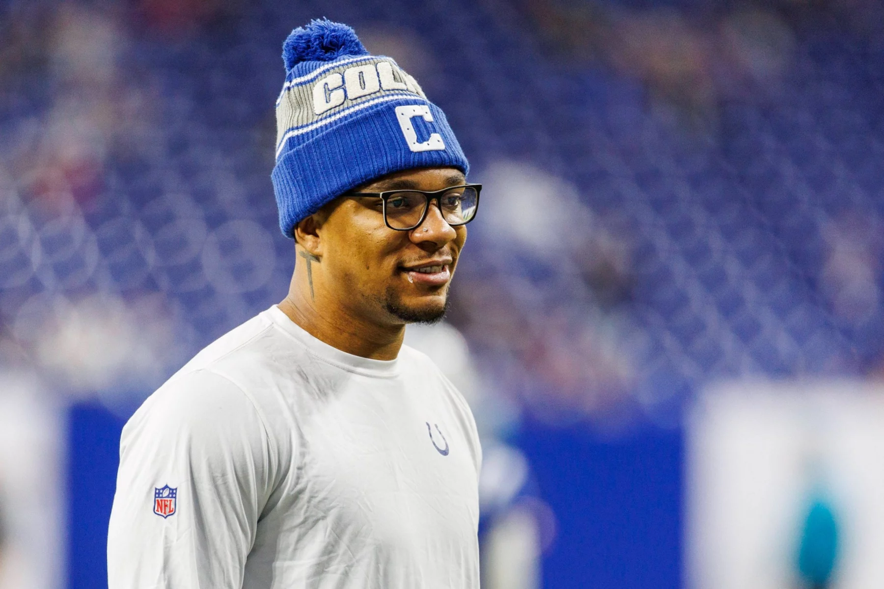 Ein lächelnder Anthony Richardson in einem weißen Hemd mit Colts-Logo und einer blauen Mütze mit der Aufschrift "COLTS" steht auf dem Spielfeld. Er trägt eine Brille und ist scharf eingestellt, während das Stadion hinter ihm nach einem spannenden Quarterback-Duell verschwimmt. Diese Beschreibung wurde mit der FootballR KI automatisch generiert.