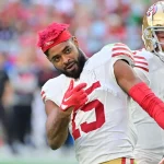 Jauan Jennings der San Francisco 49ers mit leuchtend rotem Haar und weißem Trikot mit der Nummer 15 macht während eines Spiels auf dem Spielfeld eine spielerische Geste. Ein weiterer Spieler steht in Uniform hinter ihm, während der Arm eines Schiedsrichters in den Vordergrund ragt. Diese Beschreibung wurde mit der FootballR KI automatisch generiert.