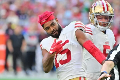 Jauan Jennings der San Francisco 49ers mit leuchtend rotem Haar und weißem Trikot mit der Nummer 15 macht während eines Spiels auf dem Spielfeld eine spielerische Geste. Ein weiterer Spieler steht in Uniform hinter ihm, während der Arm eines Schiedsrichters in den Vordergrund ragt. Diese Beschreibung wurde mit der FootballR KI automatisch generiert.