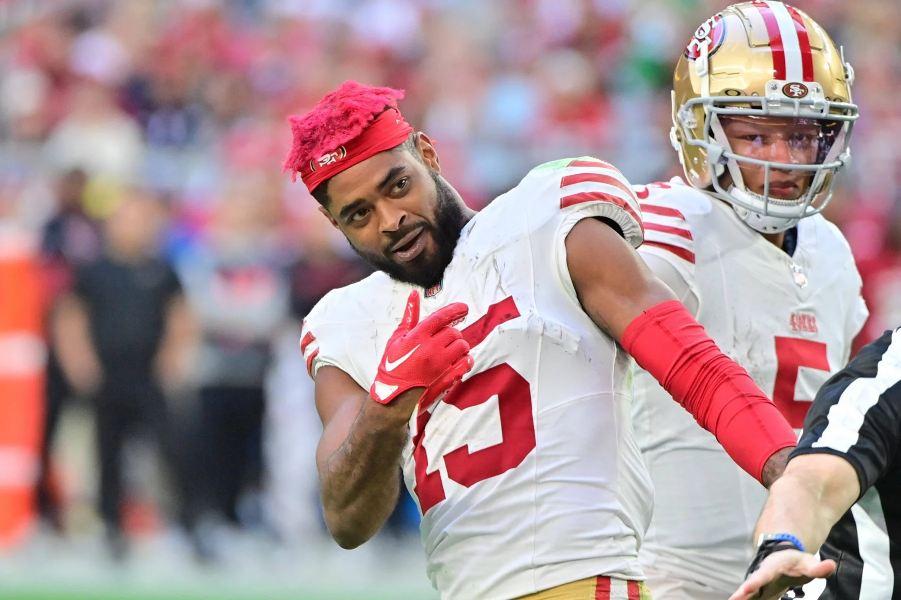 Jauan Jennings der San Francisco 49ers mit leuchtend rotem Haar und weißem Trikot mit der Nummer 15 macht während eines Spiels auf dem Spielfeld eine spielerische Geste. Ein weiterer Spieler steht in Uniform hinter ihm, während der Arm eines Schiedsrichters in den Vordergrund ragt. Diese Beschreibung wurde mit der FootballR KI automatisch generiert.