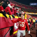 Der Spieler der Kansas City Chiefs, George Karlaftis, der die Trikotnummer 56 trägt, lächelt und gibt den eingepackten Fans, die sich über eine Absperrung im Stadion beugen, ein High-Five. Die Tribünen sind in Rot und Gelb gehalten, während im Hintergrund ein Bud Light-Schild leuchtet. Diese Beschreibung wurde mit der FootballR KI automatisch generiert.