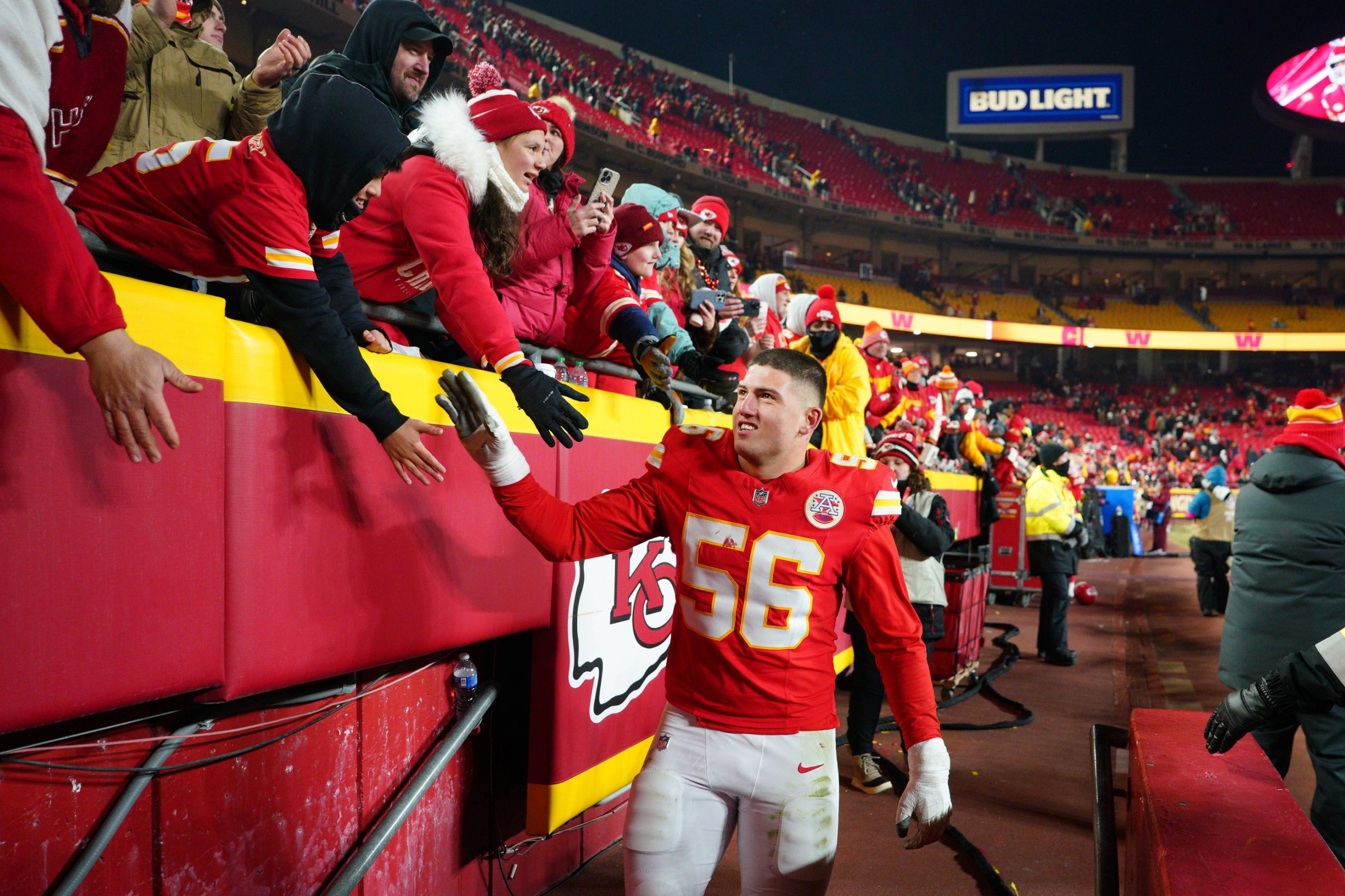Der Spieler der Kansas City Chiefs, George Karlaftis, der die Trikotnummer 56 trägt, lächelt und gibt den eingepackten Fans, die sich über eine Absperrung im Stadion beugen, ein High-Five. Die Tribünen sind in Rot und Gelb gehalten, während im Hintergrund ein Bud Light-Schild leuchtet. Diese Beschreibung wurde mit der FootballR KI automatisch generiert.